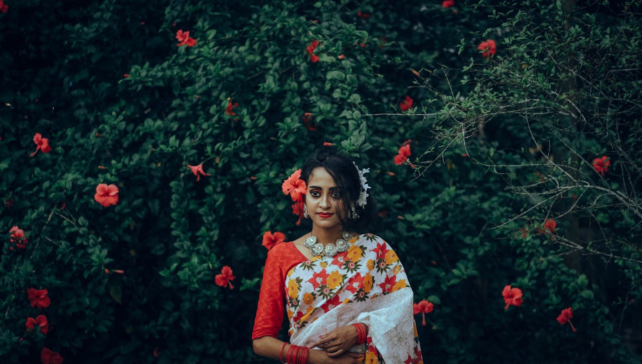 A woman in a colorful floral saree poses gracefully amidst vibrant hibiscus flowers in Faridpur, Bangladesh.